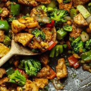 A close-up of cashew nut stir fry with tofu and vegetables in a pan. The dish includes broccoli, bell peppers, and peanuts, all tossed in a savory sauce. A wooden spoon holds a portion of the mix, highlighting the vibrant colors and texture.